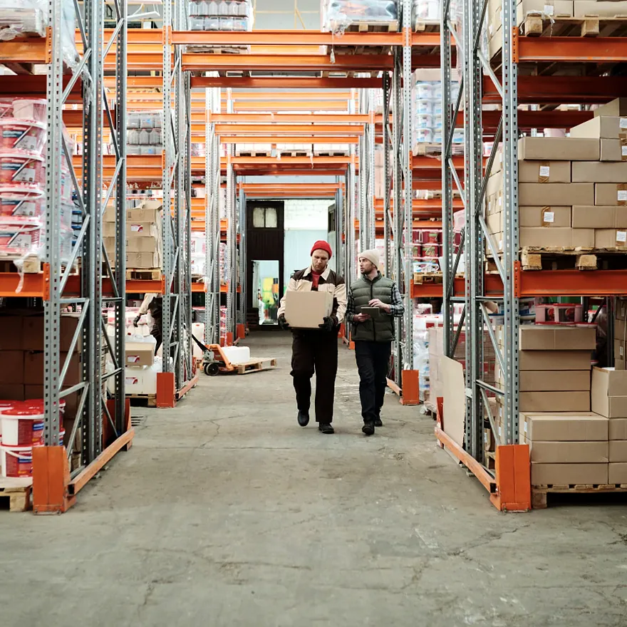Two warehouse workers walking an aisle of stocked pallet racks while moving a customer order